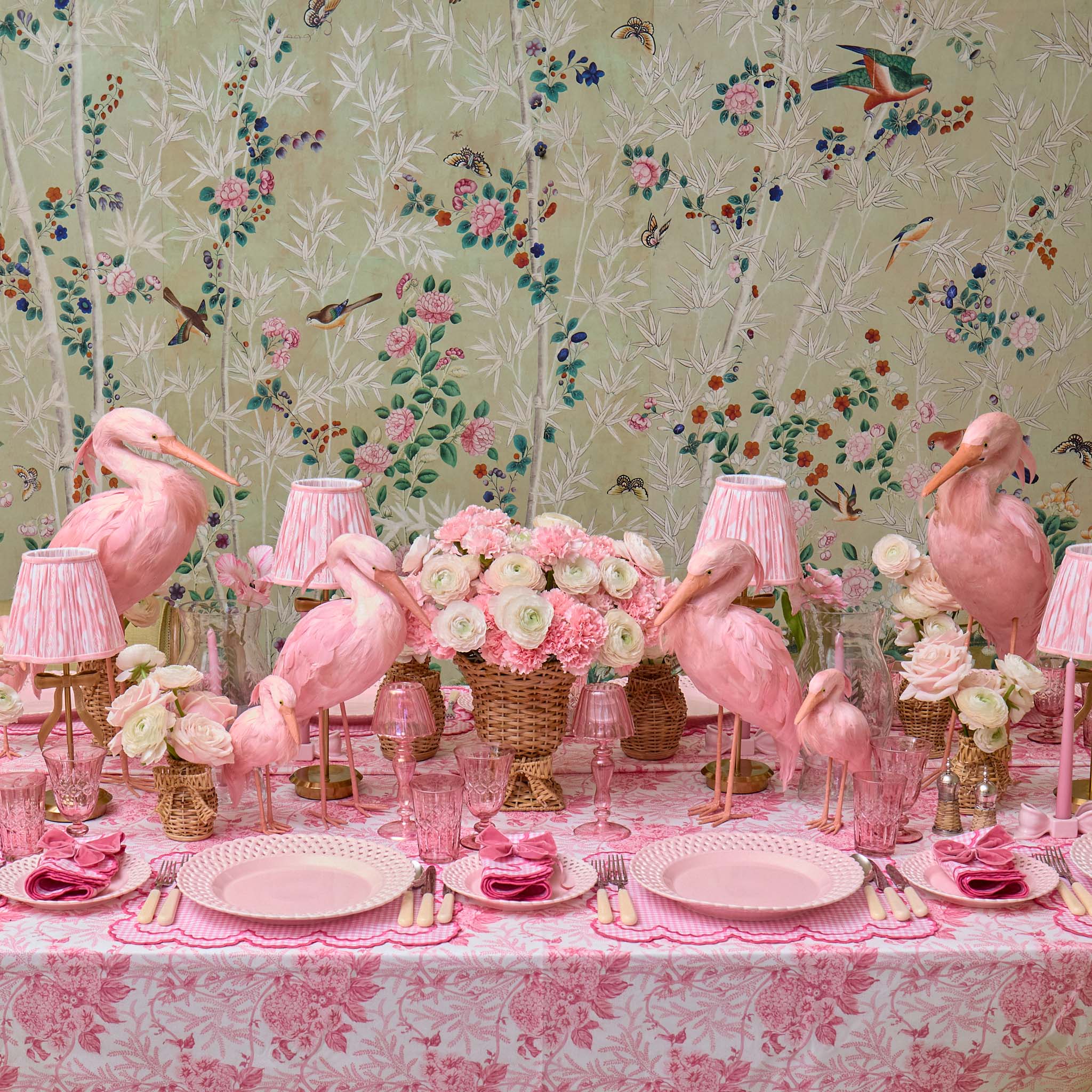 Decorative table setting with pink flamingo figurines, floral arrangements, and lamps against a floral wallpaper background.