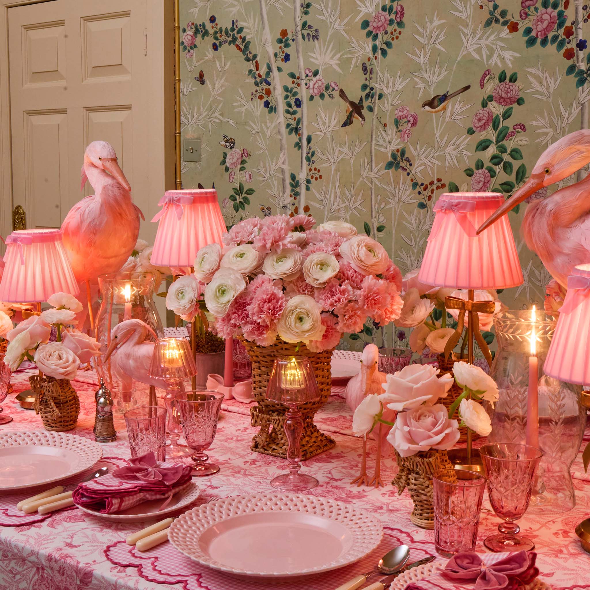 Decorative table setting with pink flamingo lamps, flowers, and tableware in a room with floral wallpaper.