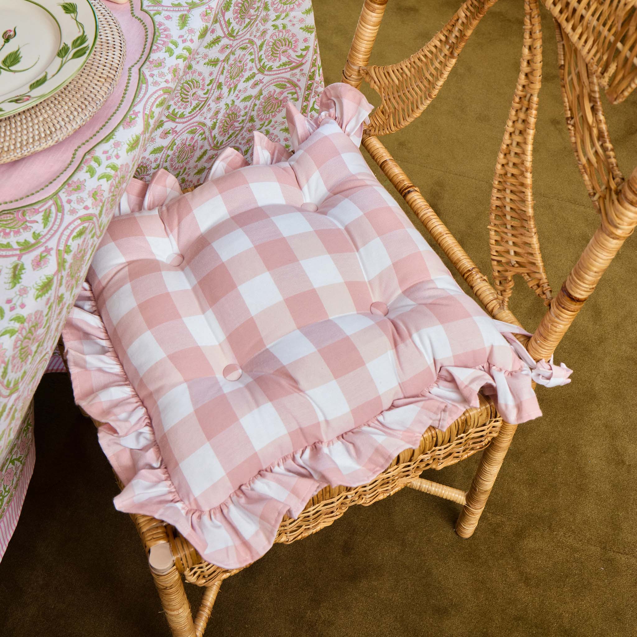 Pink checkered cushion with ruffles on a wicker chair next to a patterned tablecloth.