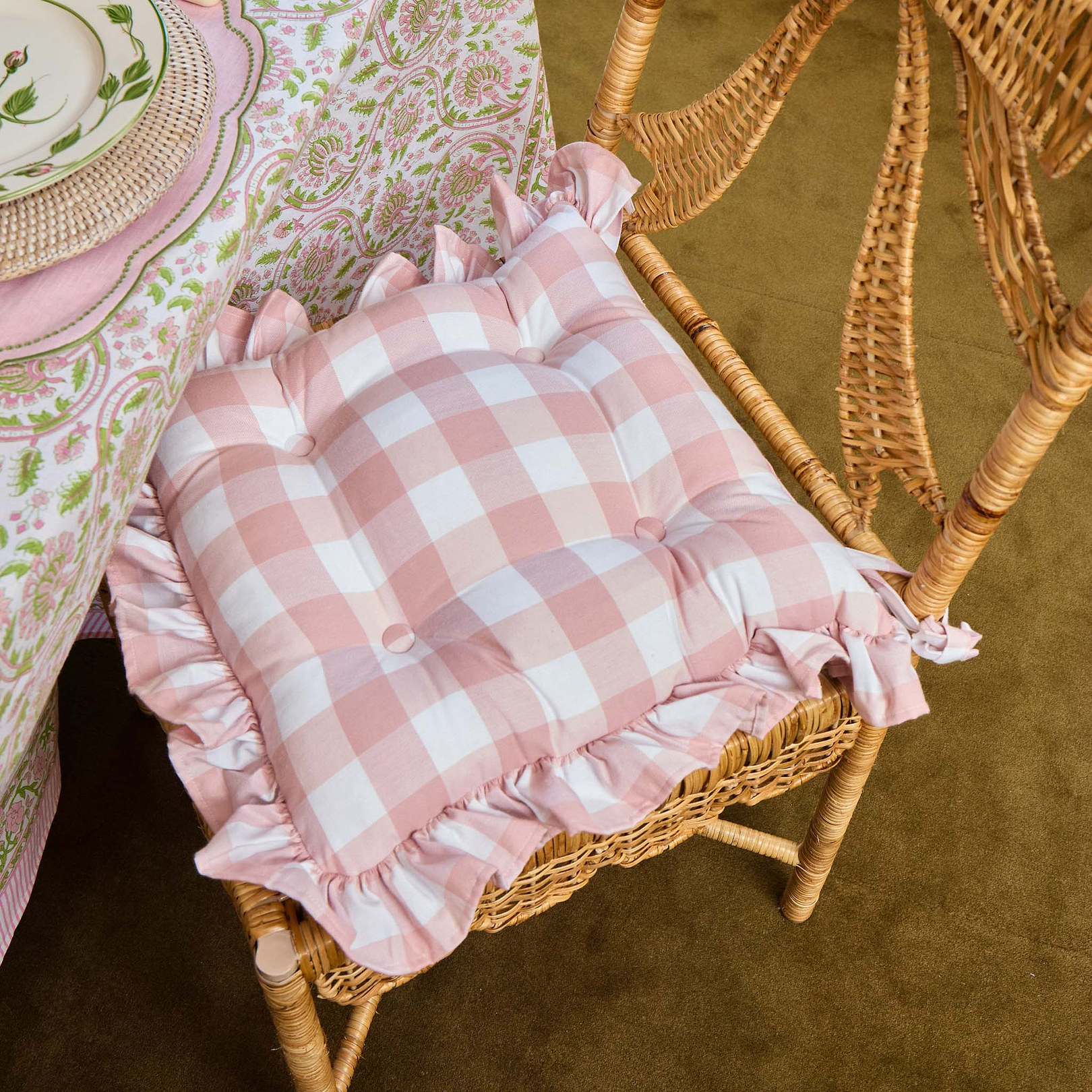 Pink checkered cushion with ruffles on a wicker chair next to a patterned tablecloth.