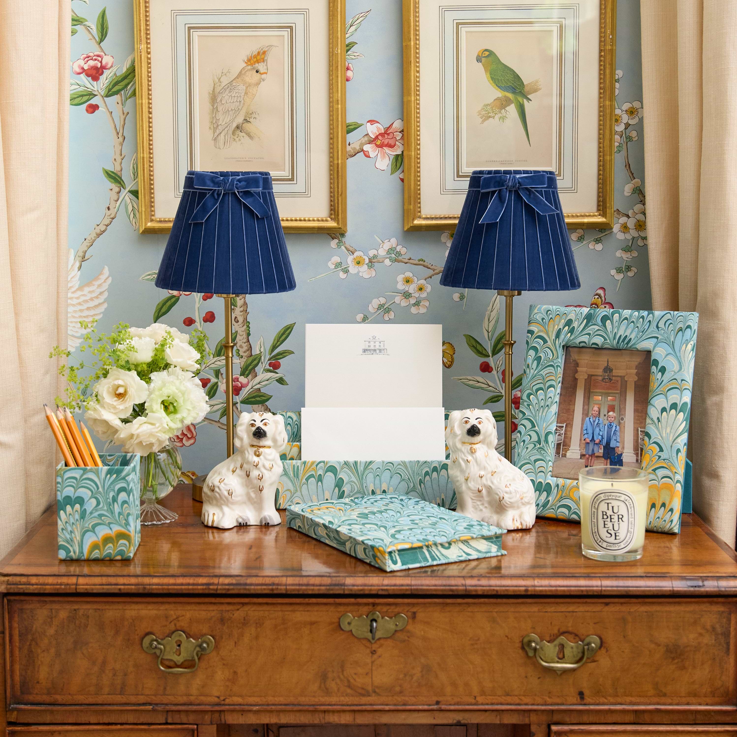 A styled home office or study scene featuring a wooden desk with a matching blue and teal patterned desk organizer, two blue pleated table lamps, ceramic dog figurines, a floral arrangement, and framed wall art against a blue floral wallpaper background.