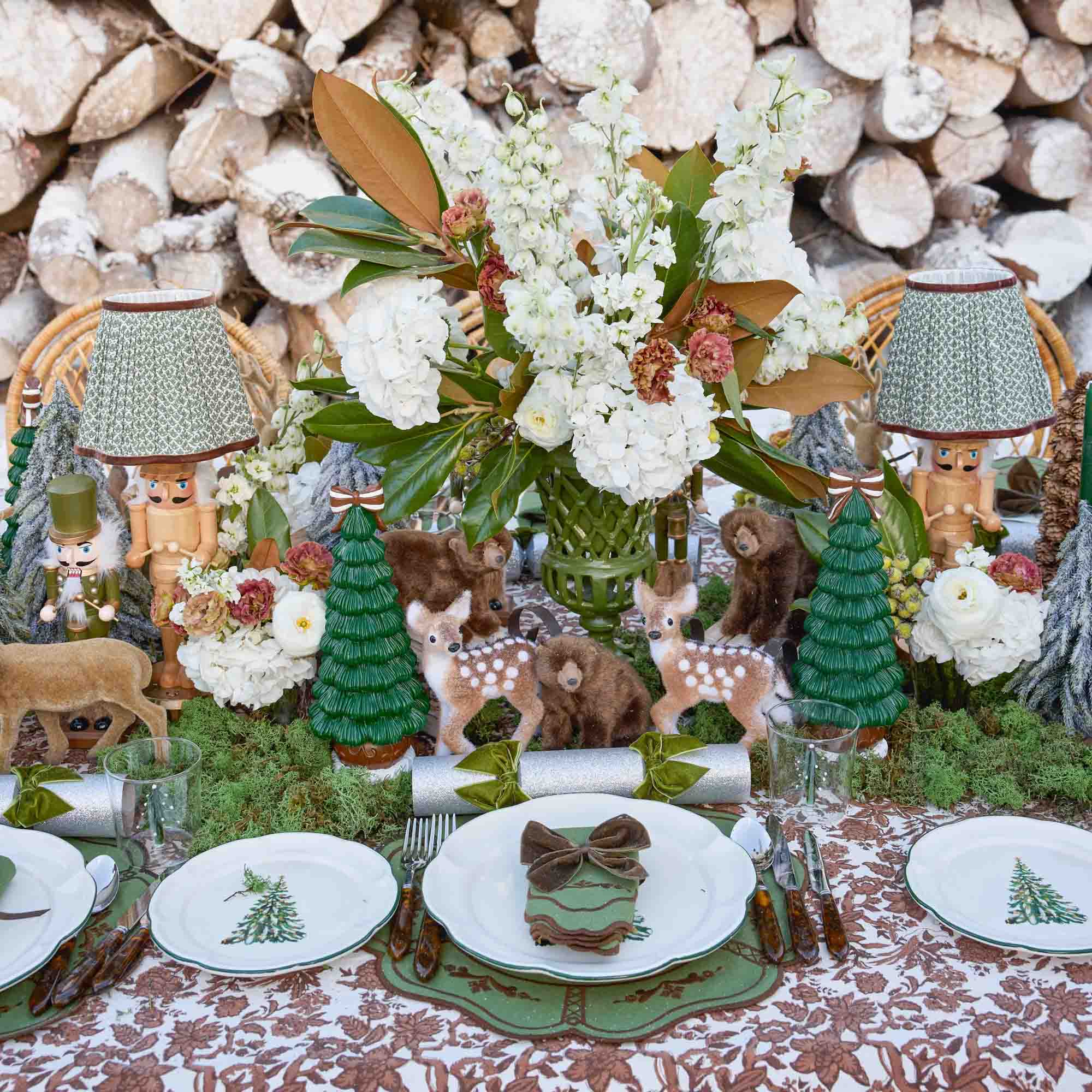 Woodland-themed table setting with floral arrangements, animal figurines, and decorative elements against a log pile background.