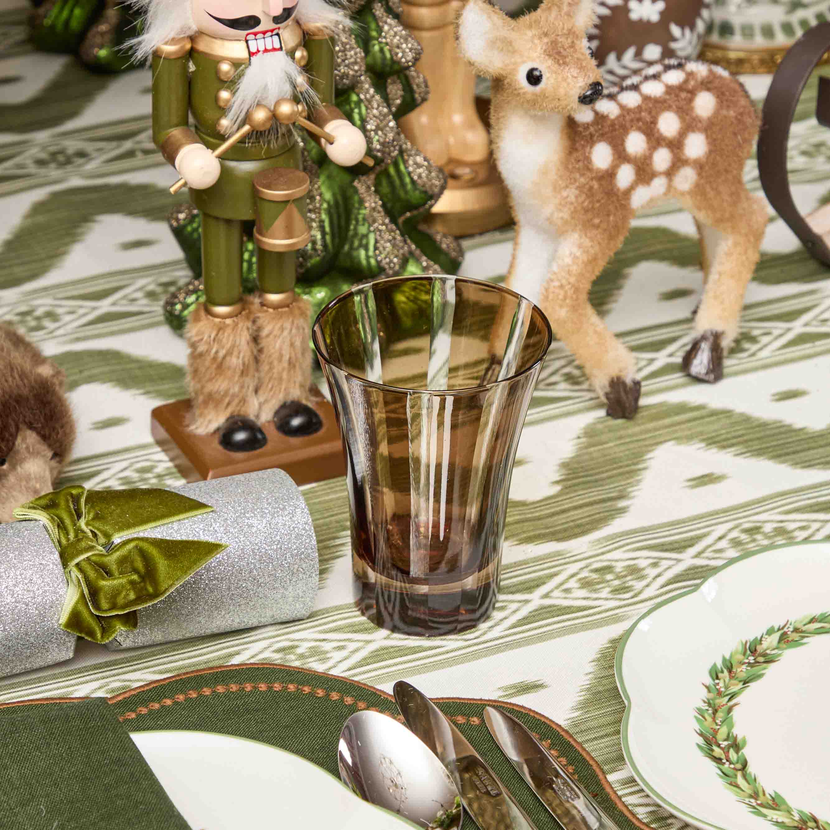 Decorative table setting with a glass, nutcracker, and deer figurine on a patterned tablecloth.