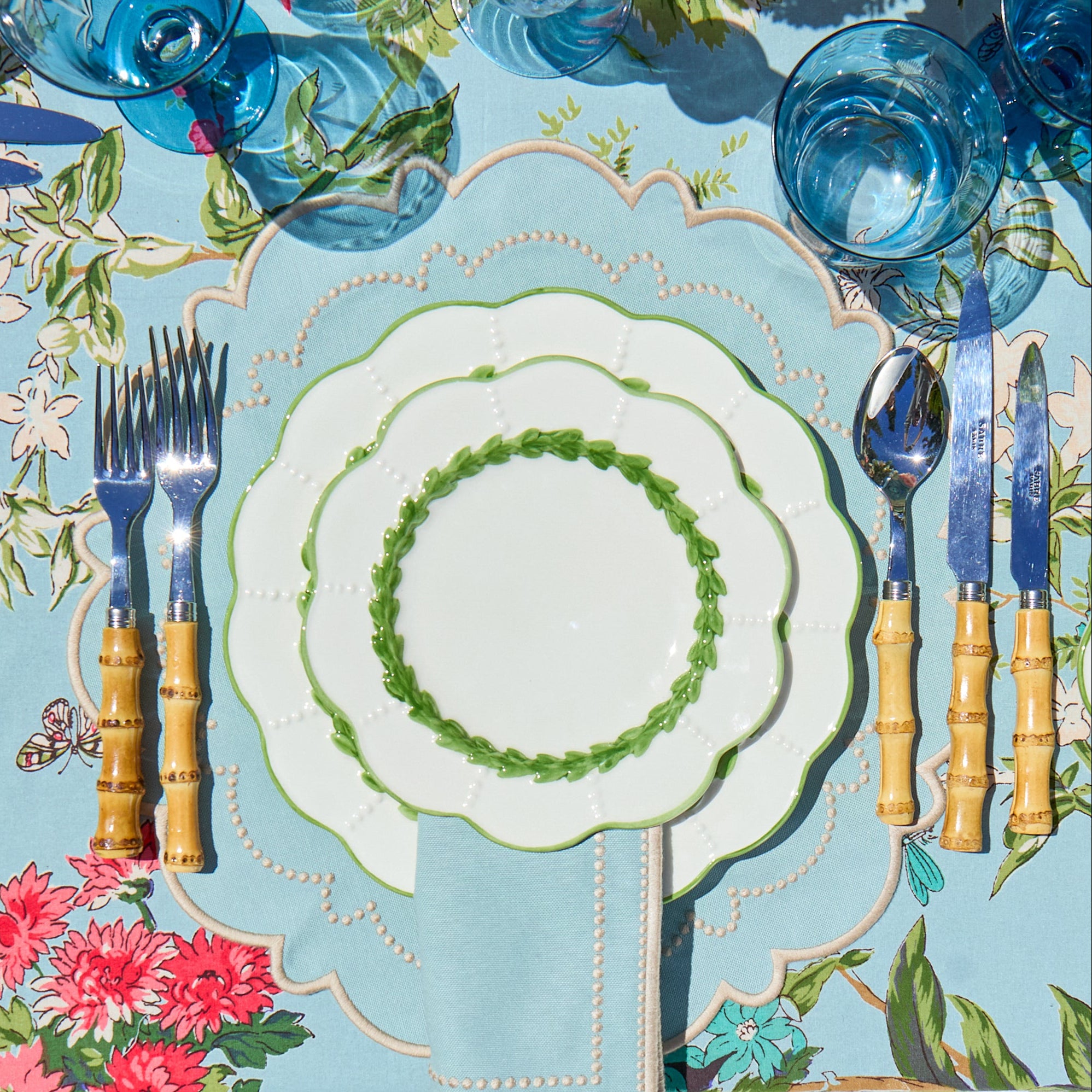 Dining table setting with floral tablecloth, white plates, blue glasses, and bamboo cutlery.
