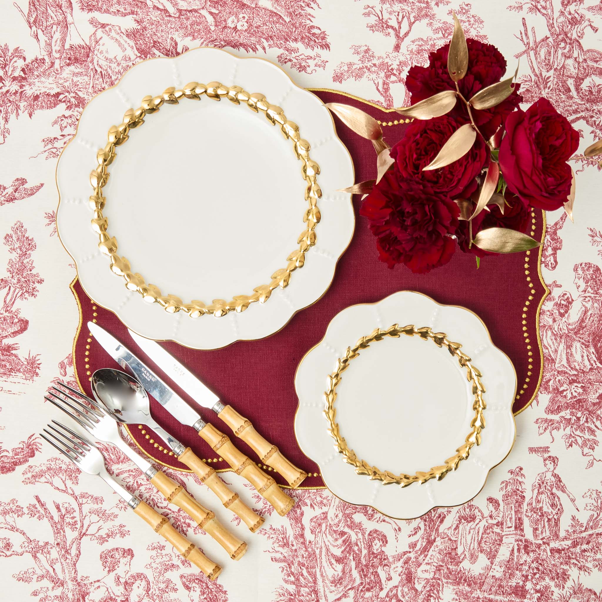 Decorative christmas  table setting with gold-rimmed plates, red roses, and silverware on a patterned tablecloth.