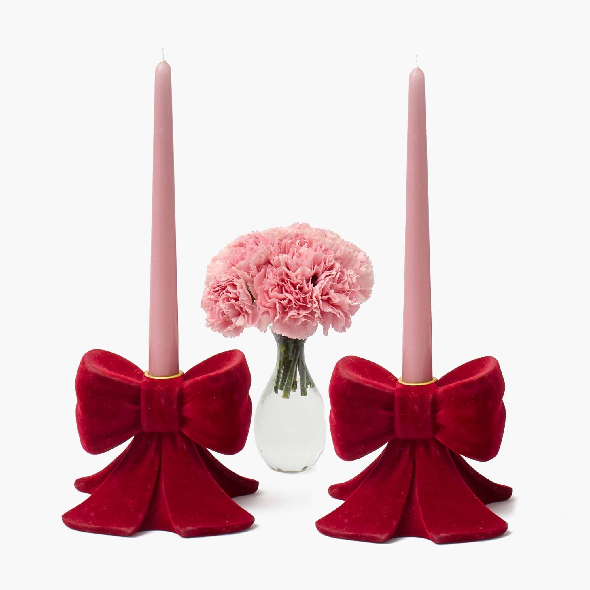 Two pink candles in red velvet bow candle holders with a vase of pink flowers in the center on a white background.