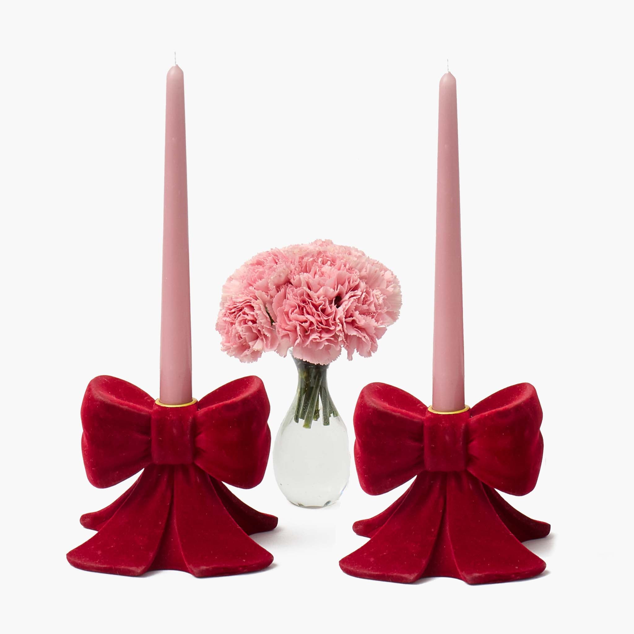 Two pink candles in red velvet bow candle holders with a vase of pink flowers in the center on a white background.