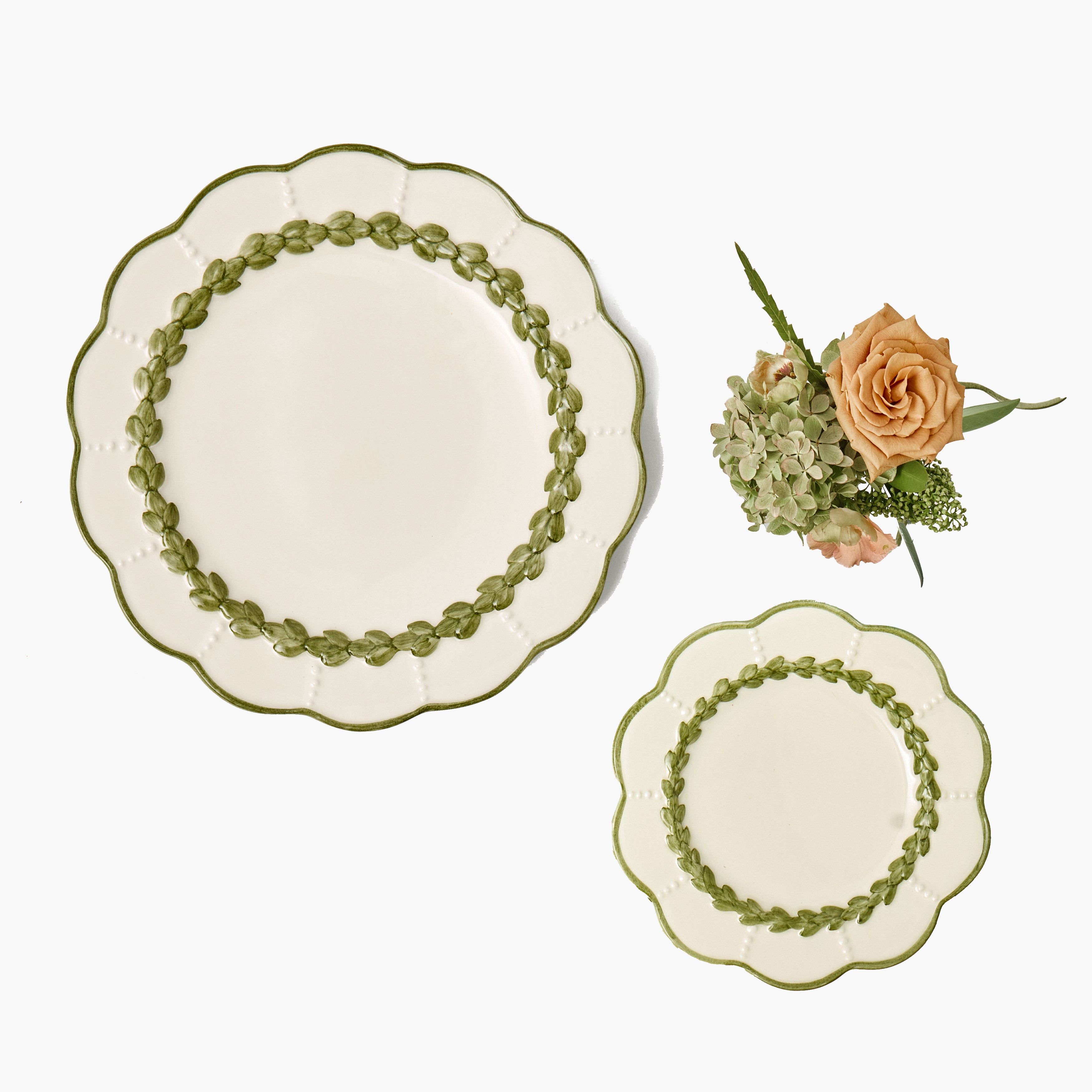 Two scalloped ceramic plates with green beaded borders, shown on a clean white background with a small floral arrangement beside them.