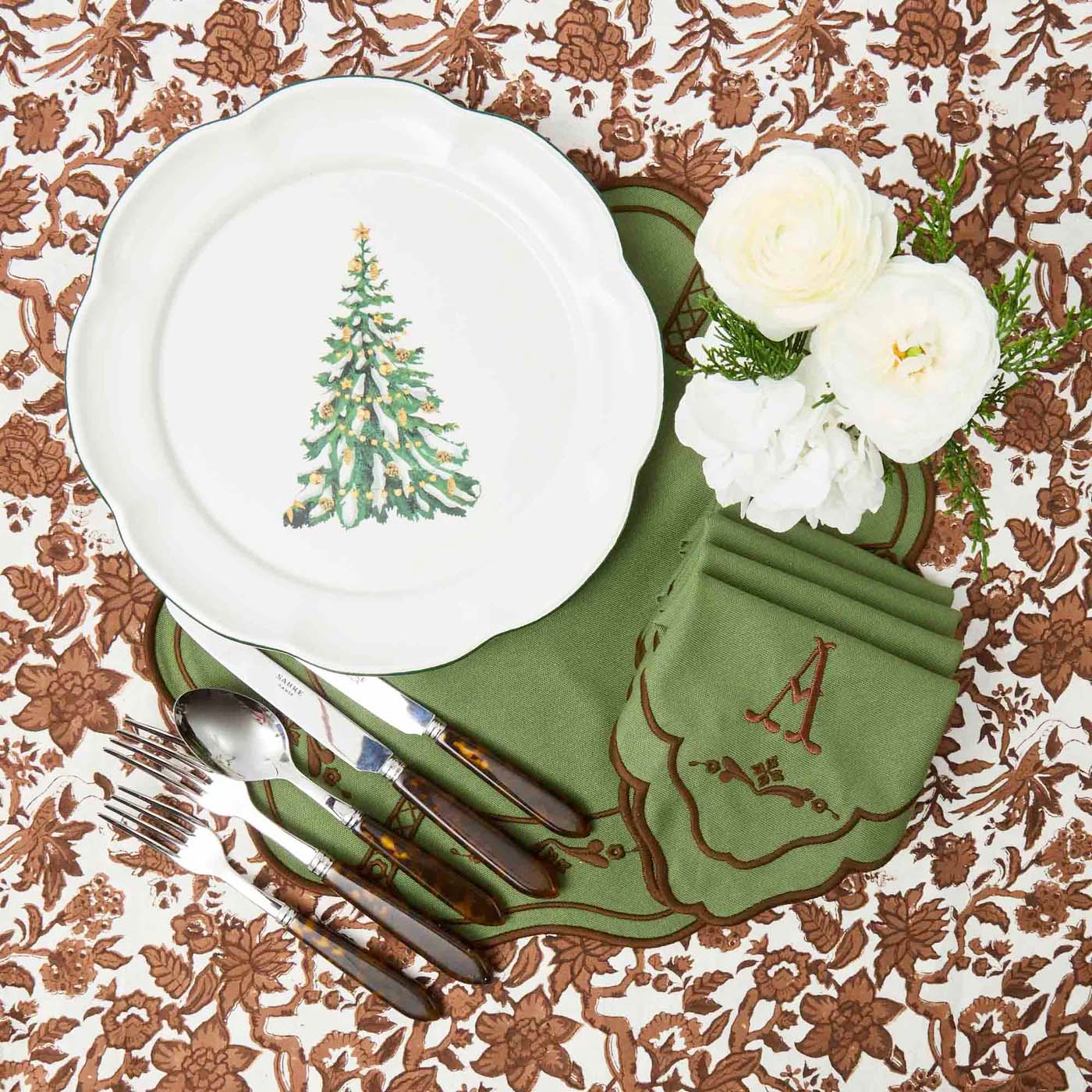 Christmas-themed table setting with a white plate featuring a green Christmas tree, green napkin, and white flowers on a patterned tablecloth.