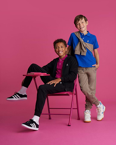 two kids on a fushia background, one is sitting and the other one is standing both smiling