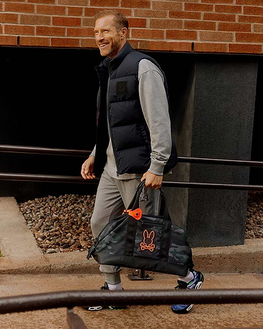 Hombre caminando con un bolso de fin de semana en la mano izquierda y vistiendo una chaqueta acolchada sin mangas de Psycho Bunny en negro con un chándal gris.