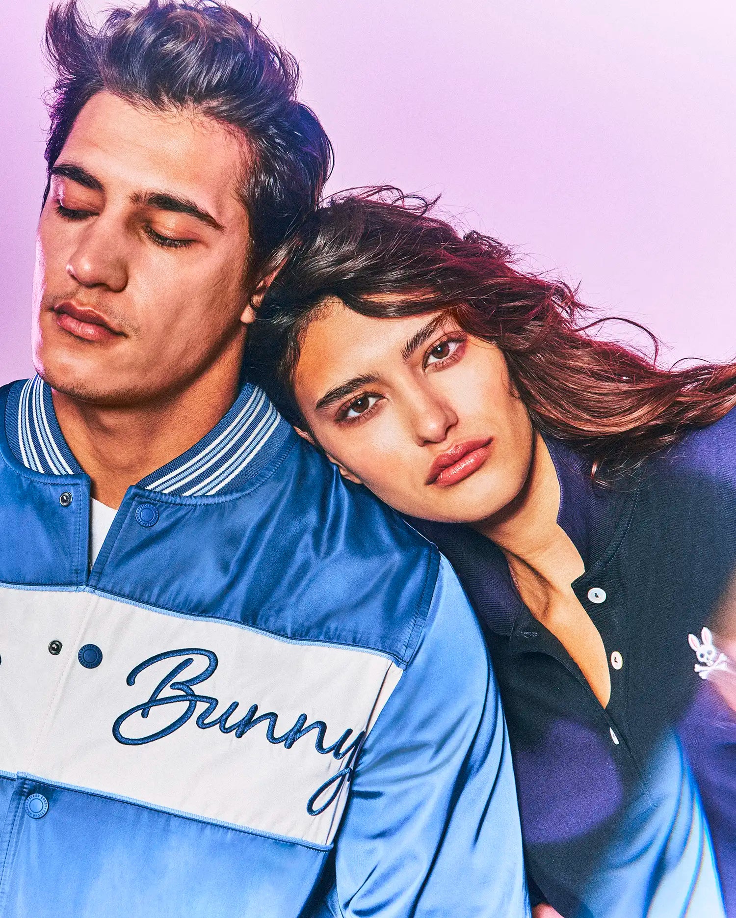 Mens Thomas Satin Bomber in two-tone blue satin with white chest stripe and embroidered Bunny script, alongside a dark blue pique polo with small white Psycho Bunny skull logo.