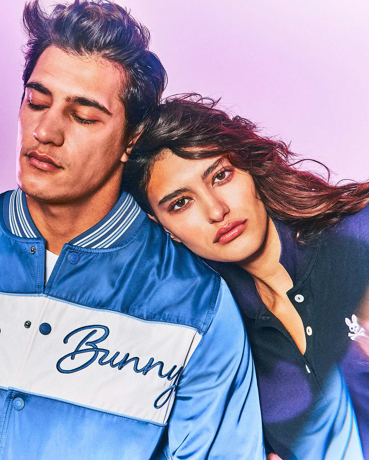 Mens Thomas Satin Bomber in two-tone blue satin with white chest stripe and embroidered Bunny script, alongside a dark blue pique polo with small white Psycho Bunny skull logo.