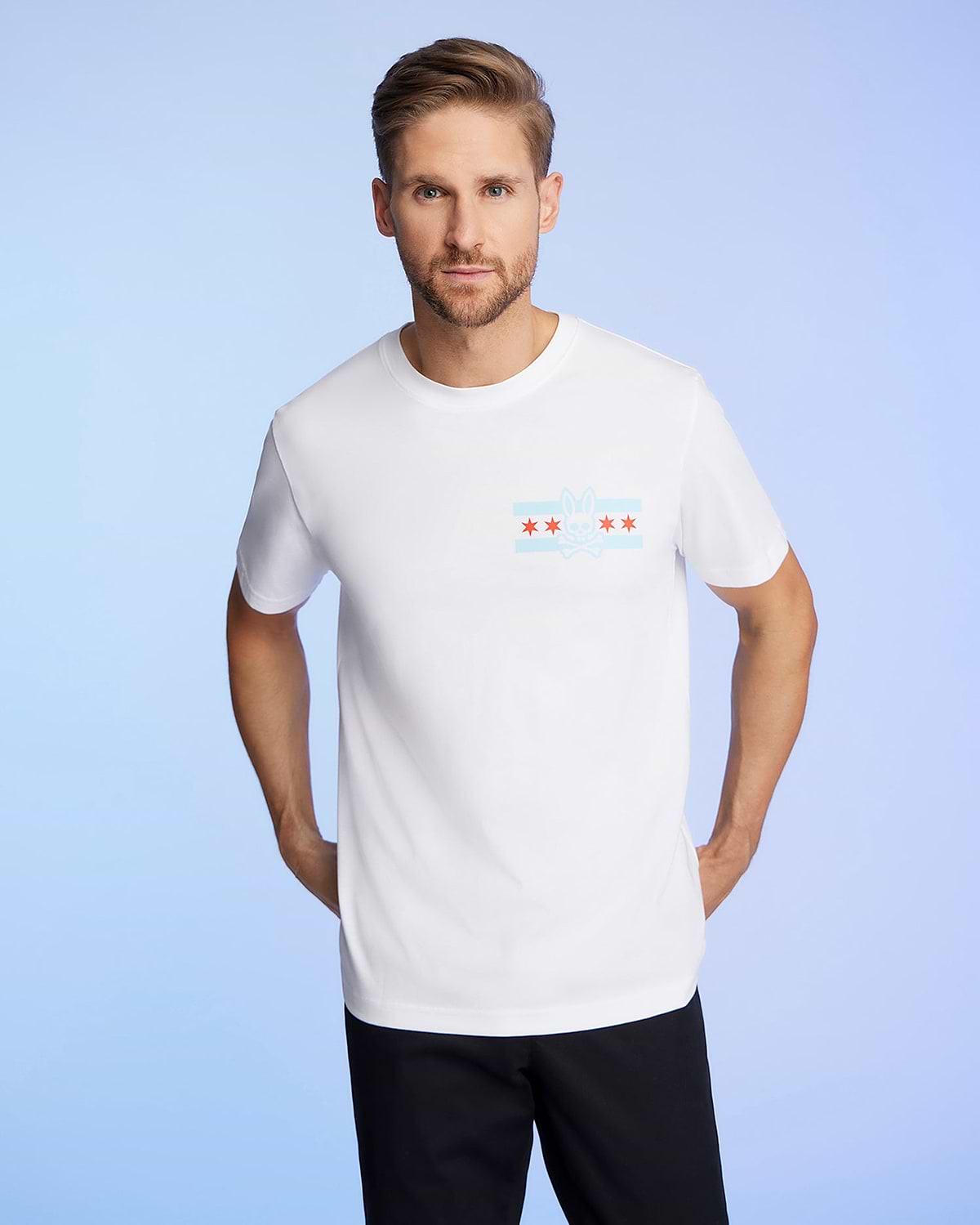 A person with light brown hair and a beard is wearing the Psycho Bunny MENS CHICAGO CITY TEE - B6U551W1PC in soft Pima cotton. The shirt has a graphic design of red stars and blue stripes on the chest against a light blue gradient background.