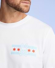 A man wears the Psycho Bunny MENS CHICAGO CITY TEE - B6U551W1PC, a white Pima cotton shirt featuring a blue and red rabbit skull and crossbones graphic with red stars on the chest, set against a light blue background for a vibrant look.