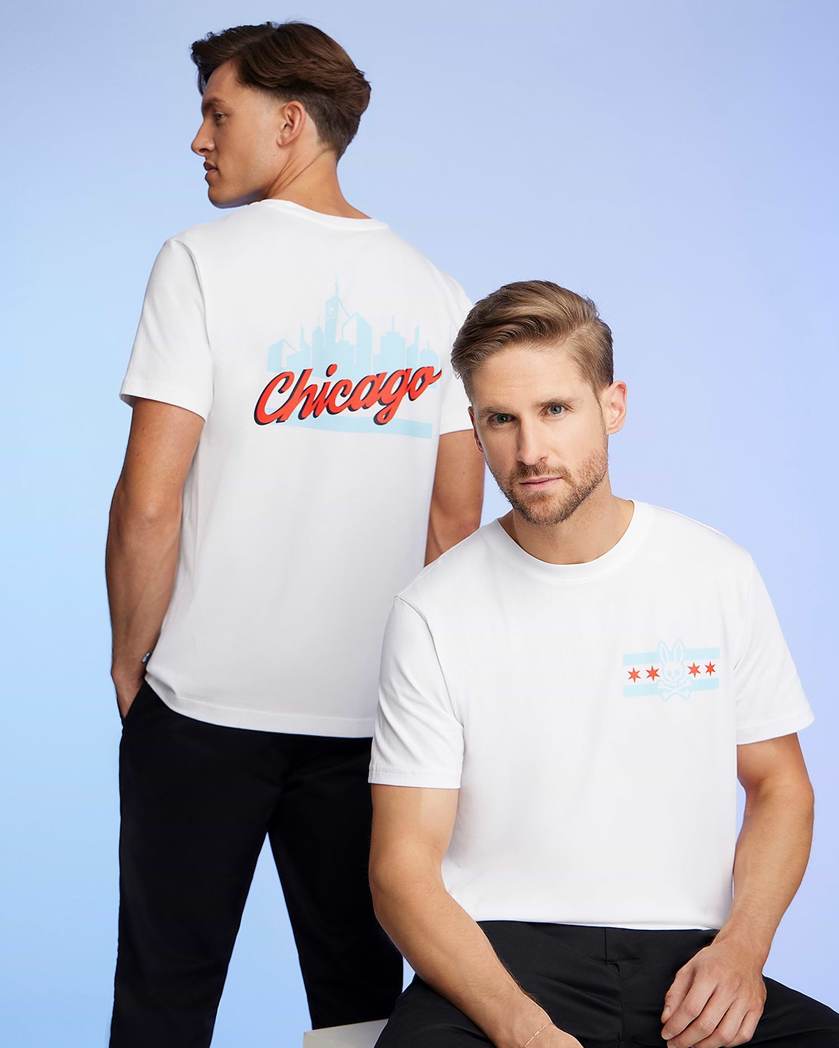 Two men wear Chicago-themed Psycho Bunny tees made from soft Pima cotton. One displays "Chicago" and the skyline on his back, while the other features the city's flag and skyline on the front. The signature Bunny logo adorns each shirt against a calming blue background.