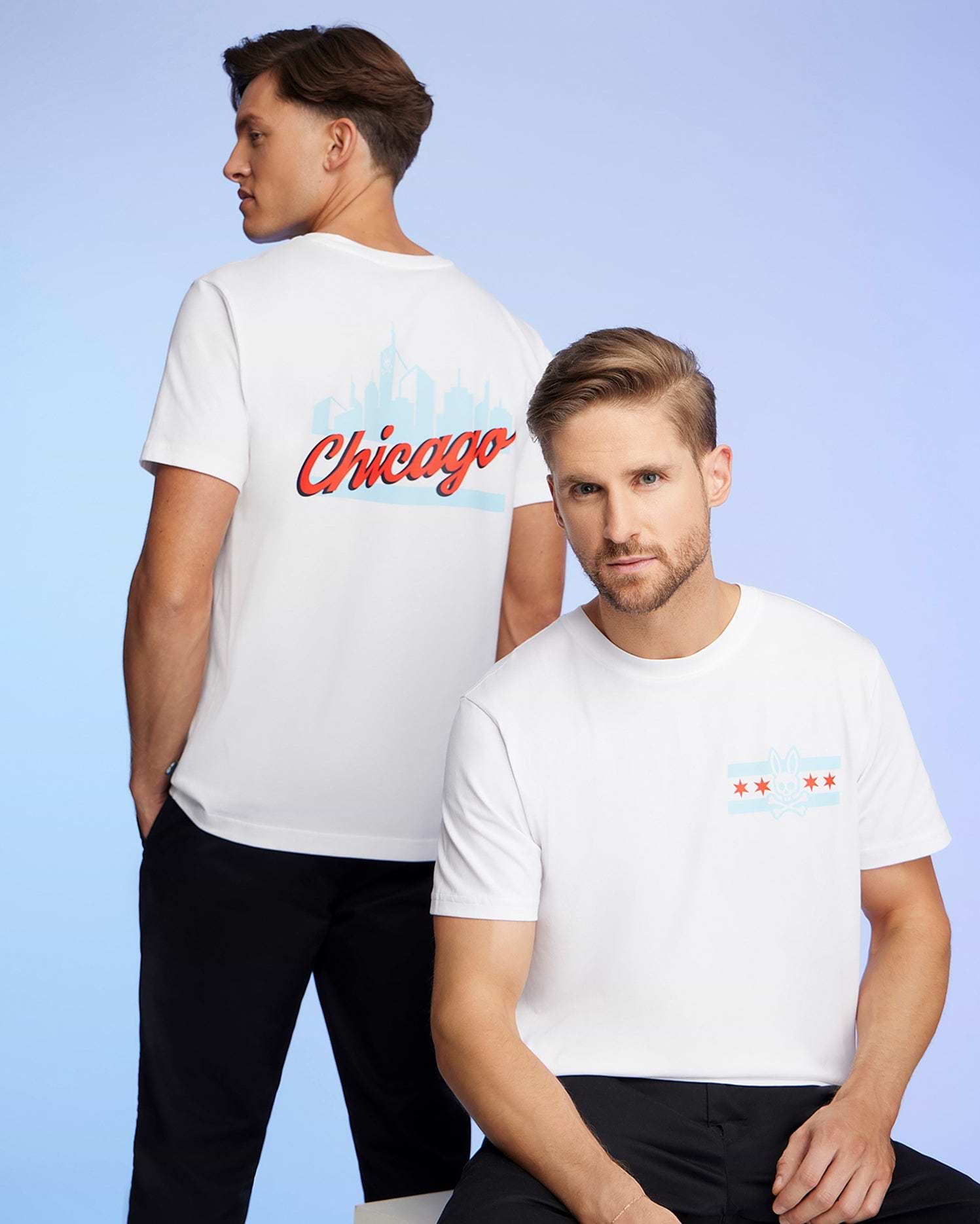 Two men wear Chicago-themed Psycho Bunny tees made from soft Pima cotton. One displays "Chicago" and the skyline on his back, while the other features the city's flag and skyline on the front. The signature Bunny logo adorns each shirt against a calming blue background.