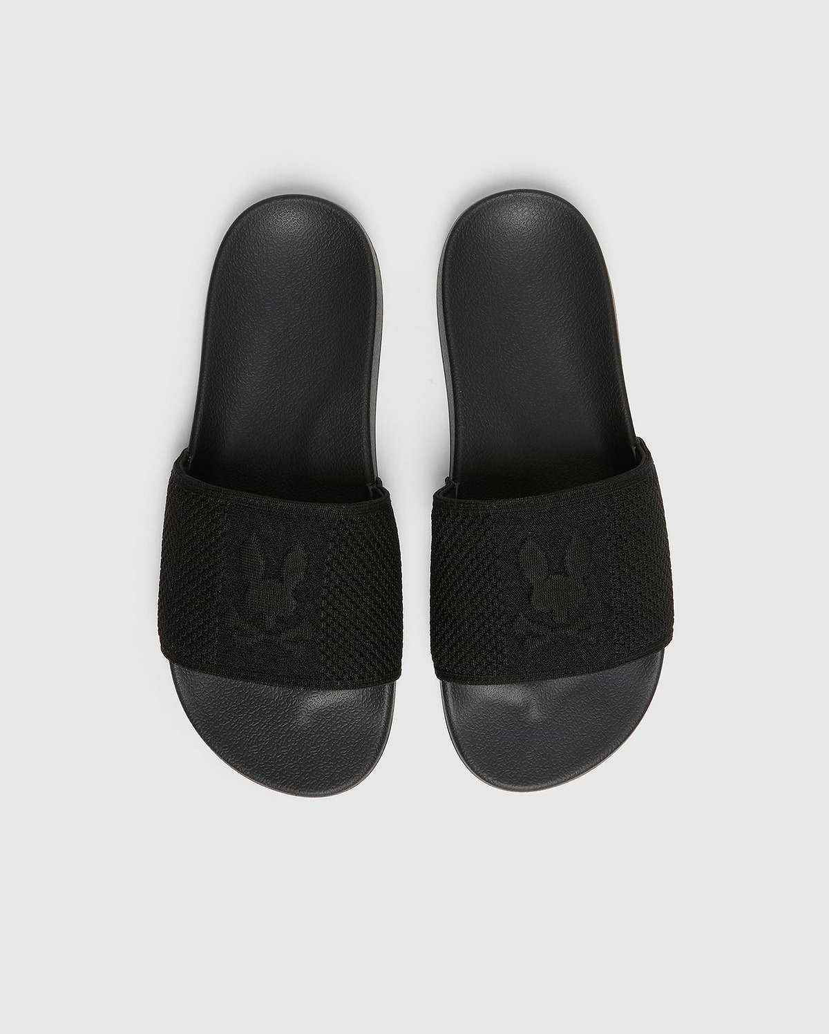 A pair of black Psycho Bunny KIDS KNIT SLIDES - B0A377F200, featuring a textured strap with an embroidered stylized rabbit head logo and a molded footbed for comfort, are placed side by side on a light gray background.