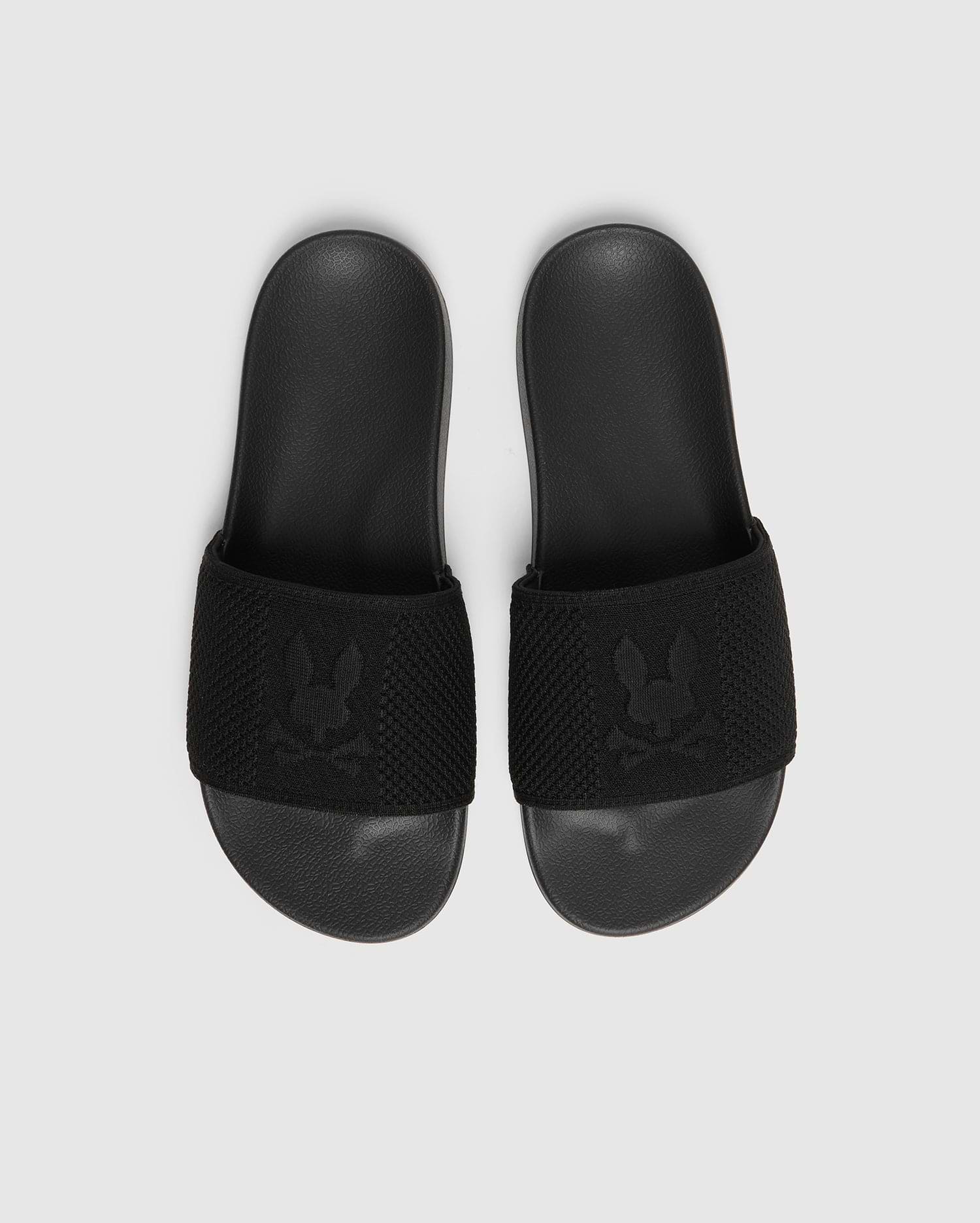 A pair of black Psycho Bunny KIDS KNIT SLIDES - B0A377F200, featuring a textured strap with an embroidered stylized rabbit head logo and a molded footbed for comfort, are placed side by side on a light gray background.