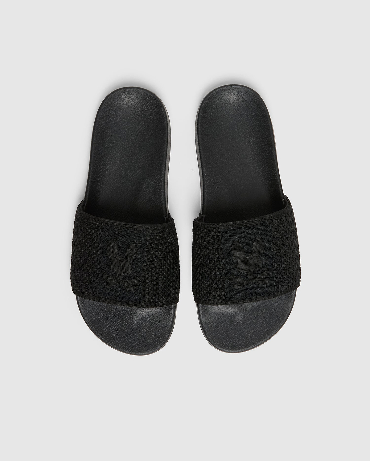 A pair of black Psycho Bunny KIDS KNIT SLIDES - B0A377F200, featuring a textured strap with an embroidered stylized rabbit head logo and a molded footbed for comfort, are placed side by side on a light gray background.