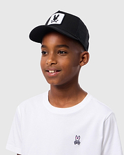 A boy wears the Psycho Bunny KIDS BUNNY PATCH TRUCKER CAP - B0A965C200 with breathable mesh paneling and a black bunny logo patch, paired with a white T-shirt featuring a small pink bunny, standing against a light gray background.