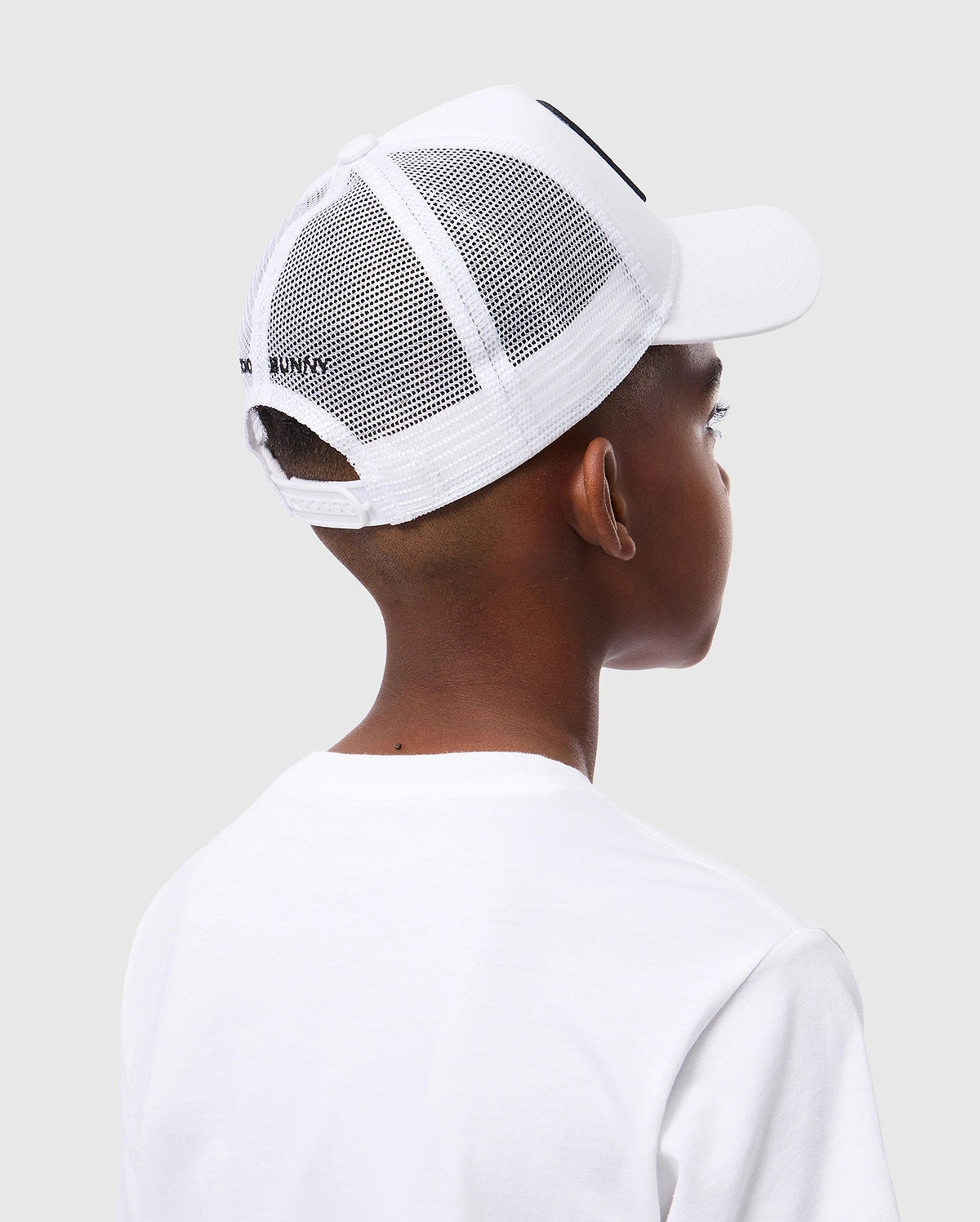 A person in a white t-shirt and a Psycho Bunny KIDS BUNNY PATCH TRUCKER CAP - B0A965C200 is seen from the back against a light grey background. The cap has the recognizable Bunny logo patch, with short hair visible beneath it.