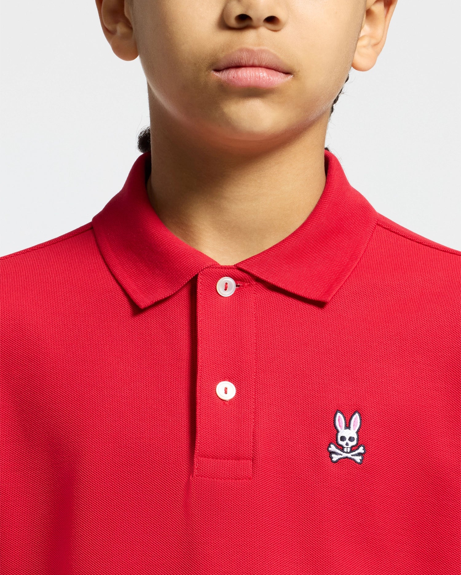 Vibrant red kids classic pique polo shirt featuring a two-button placket with white buttons, a ribbed collar, and a small, embroidered white, black, and pink Psycho Bunny skull logo on the left chest.