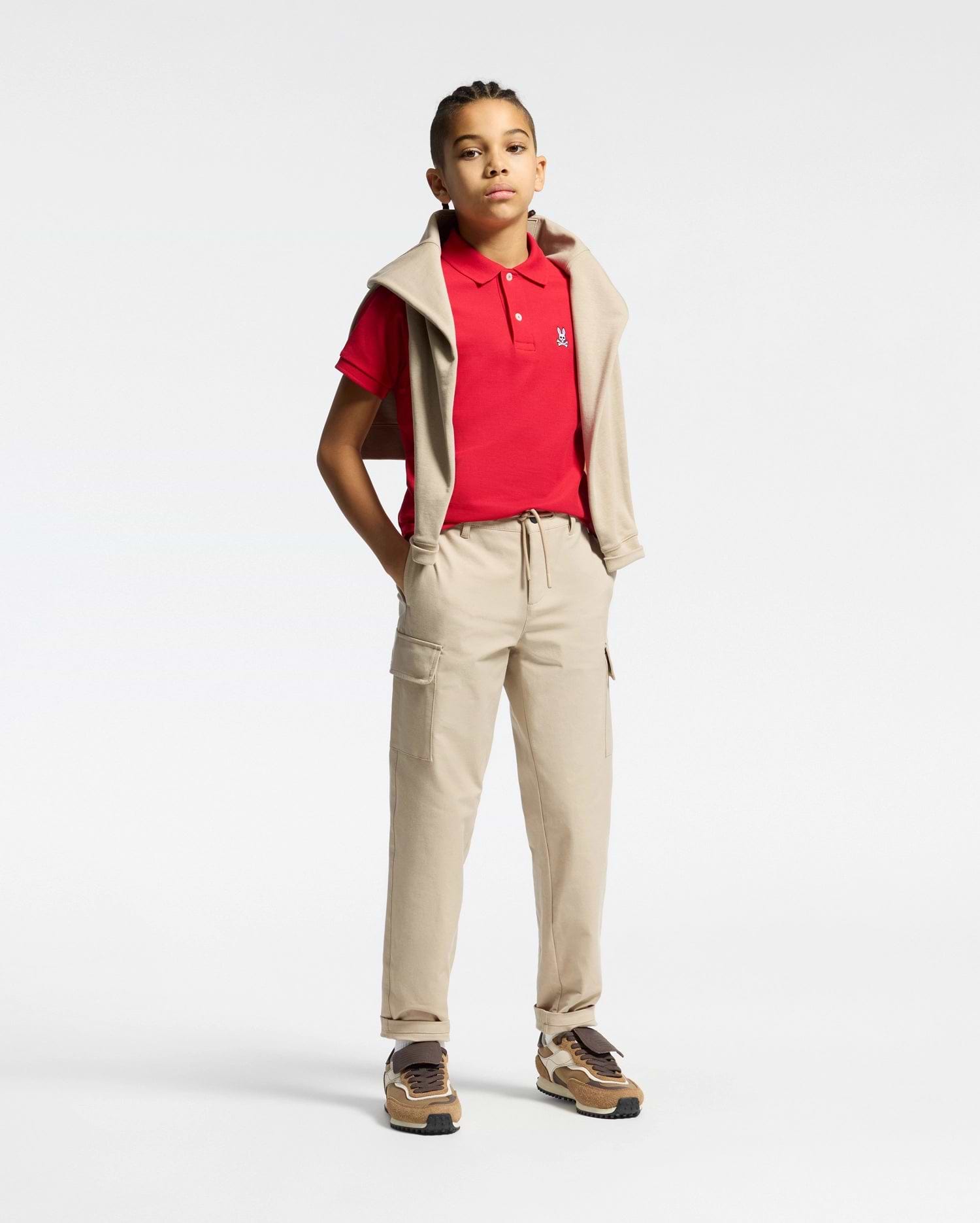 Red kids classic pique polo shirt with a classic fit, white two-button placket, and small embroidered white Psycho Bunny skull logo on the left chest, styled with beige cargo pants and a matching sweatshirt.