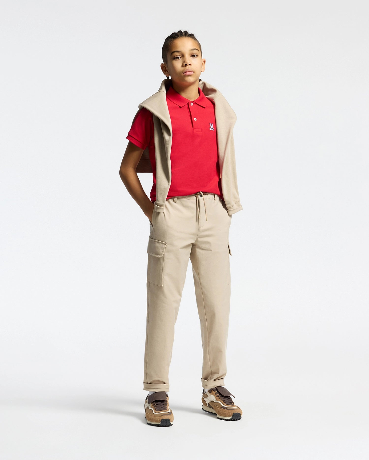 Red kids classic pique polo shirt with a classic fit, white two-button placket, and small embroidered white Psycho Bunny skull logo on the left chest, styled with beige cargo pants and a matching sweatshirt.