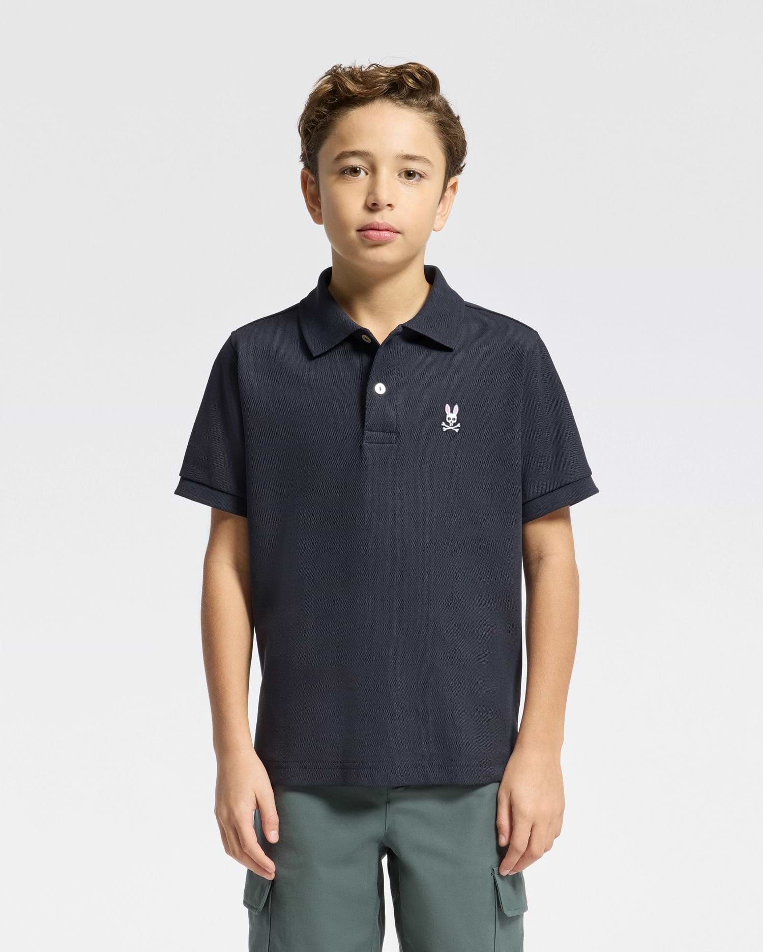 A boy with short brown hair wears the Psycho Bunny KIDS CLASSIC PIQUE POLO SHIRT (B0K001CRPC) featuring a small embroidered logo and green pants, standing against a plain light background.