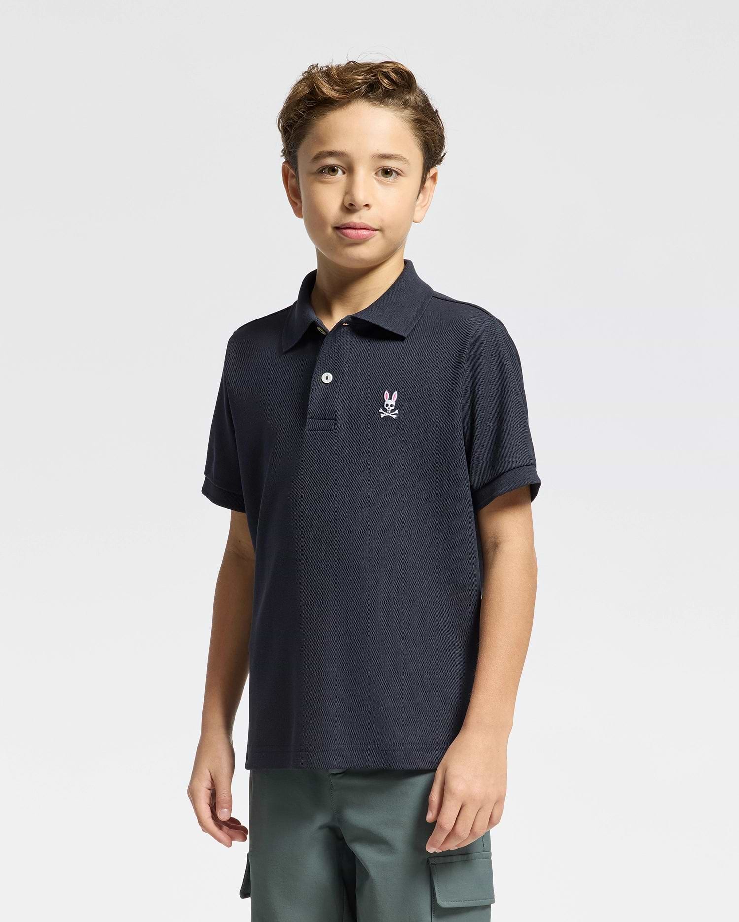 A young boy with short brown hair wears a Psycho Bunny KIDS CLASSIC PIQUE POLO SHIRT (B0K001CRPC) in navy blue Pima cotton with a white chest logo and green cargo shorts, standing against a plain light background.