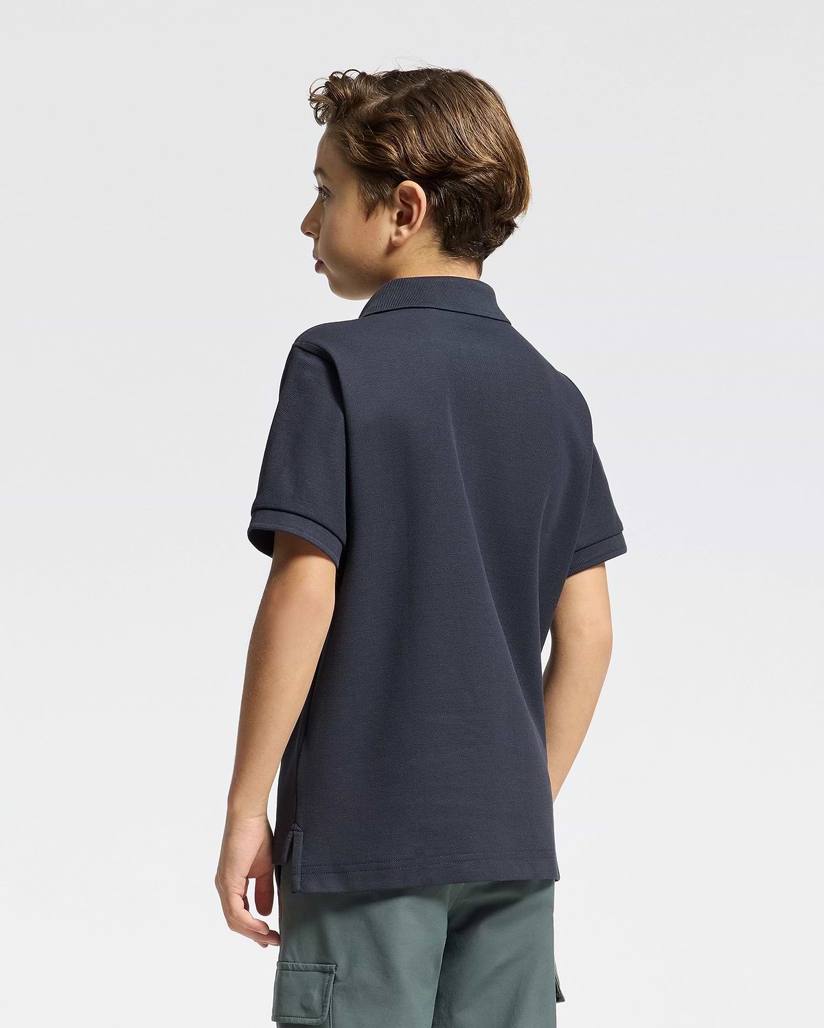 A young boy with short brown hair, seen from behind, is wearing a Psycho Bunny KIDS CLASSIC PIQUE POLO SHIRT - B0K001CRPC in dark navy and green cargo shorts, set against a plain light background.