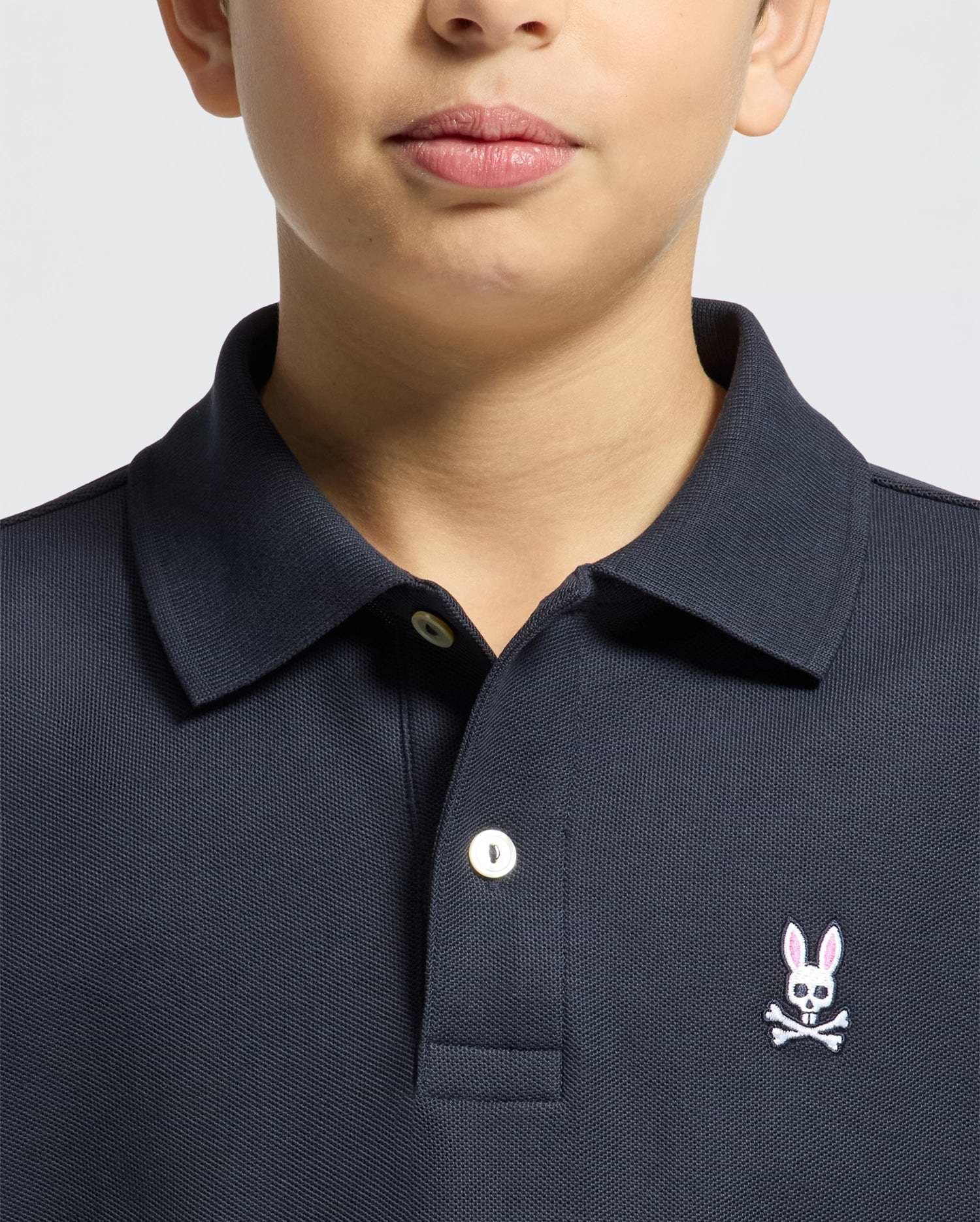 A child wears a Psycho Bunny KIDS CLASSIC PIQUE POLO SHIRT (B0K001CRPC) in dark Pima cotton, with a white button and pink embroidered bunny with crossbones on the left chest; only the lower face and upper torso are shown.