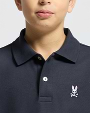 A child wears a Psycho Bunny KIDS CLASSIC PIQUE POLO SHIRT (B0K001CRPC) in dark Pima cotton, with a white button and pink embroidered bunny with crossbones on the left chest; only the lower face and upper torso are shown.