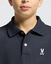 A child wears a Psycho Bunny KIDS CLASSIC PIQUE POLO SHIRT (B0K001CRPC) in dark Pima cotton, with a white button and pink embroidered bunny with crossbones on the left chest; only the lower face and upper torso are shown.