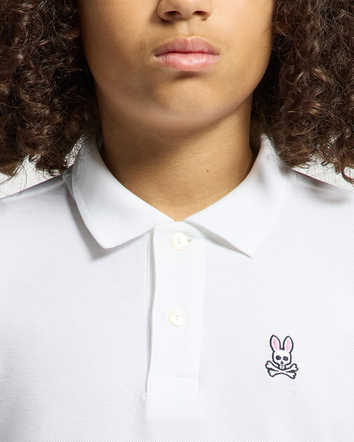 A person with curly hair is wearing a Psycho Bunny KIDS CLASSIC PIQUE POLO SHIRT in soft Pima cotton, adorned with a small pink bunny logo and crossbones on the left side. The image shows their upper torso and face, excluding the eyes.