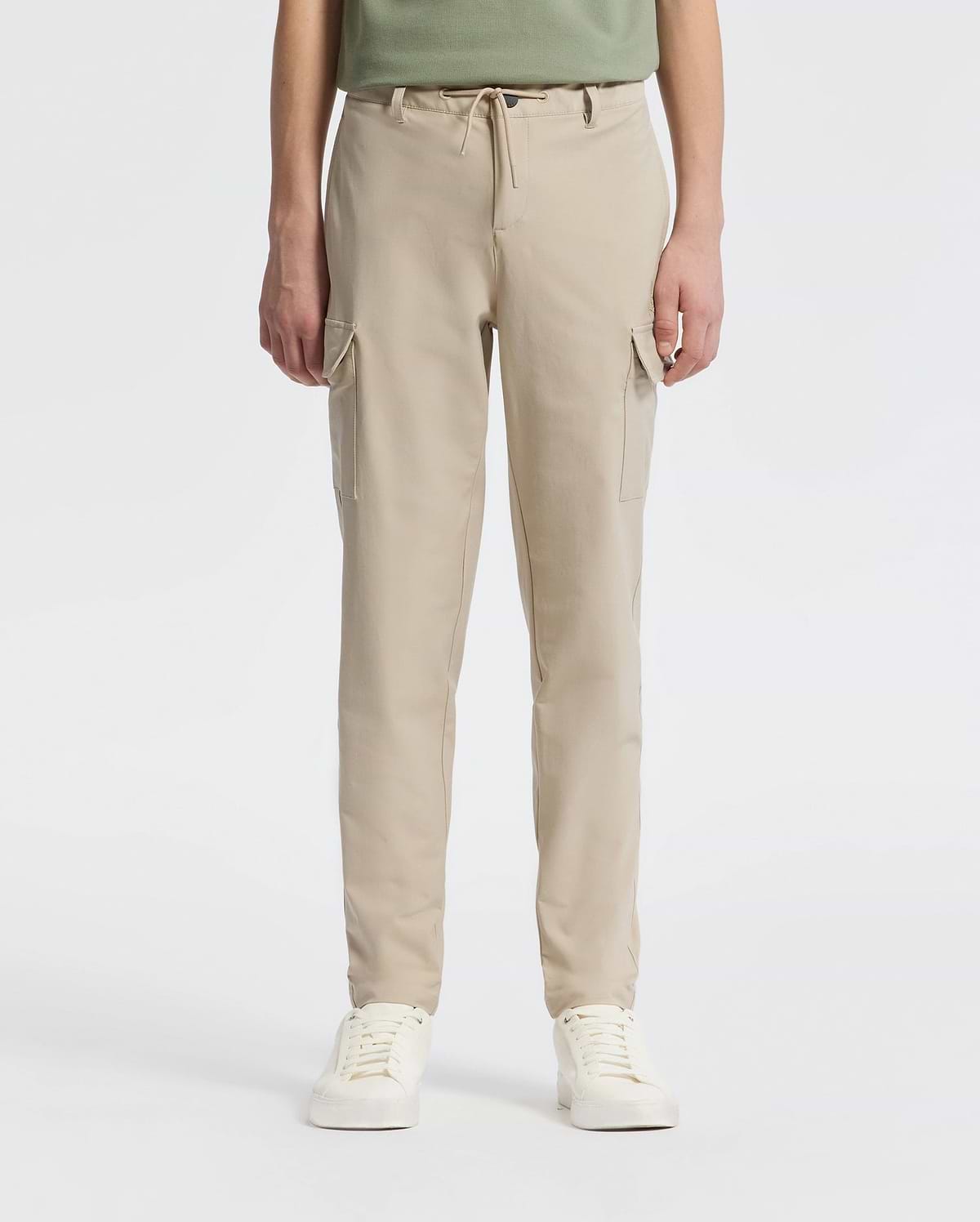A person stands against a plain backdrop wearing Psycho Bunny’s KIDS GREEN VALLEY CARGO PANT - B0P199E200, featuring beige drawstring waist and multiple pockets, paired with white sneakers. The upper body is partially visible.
