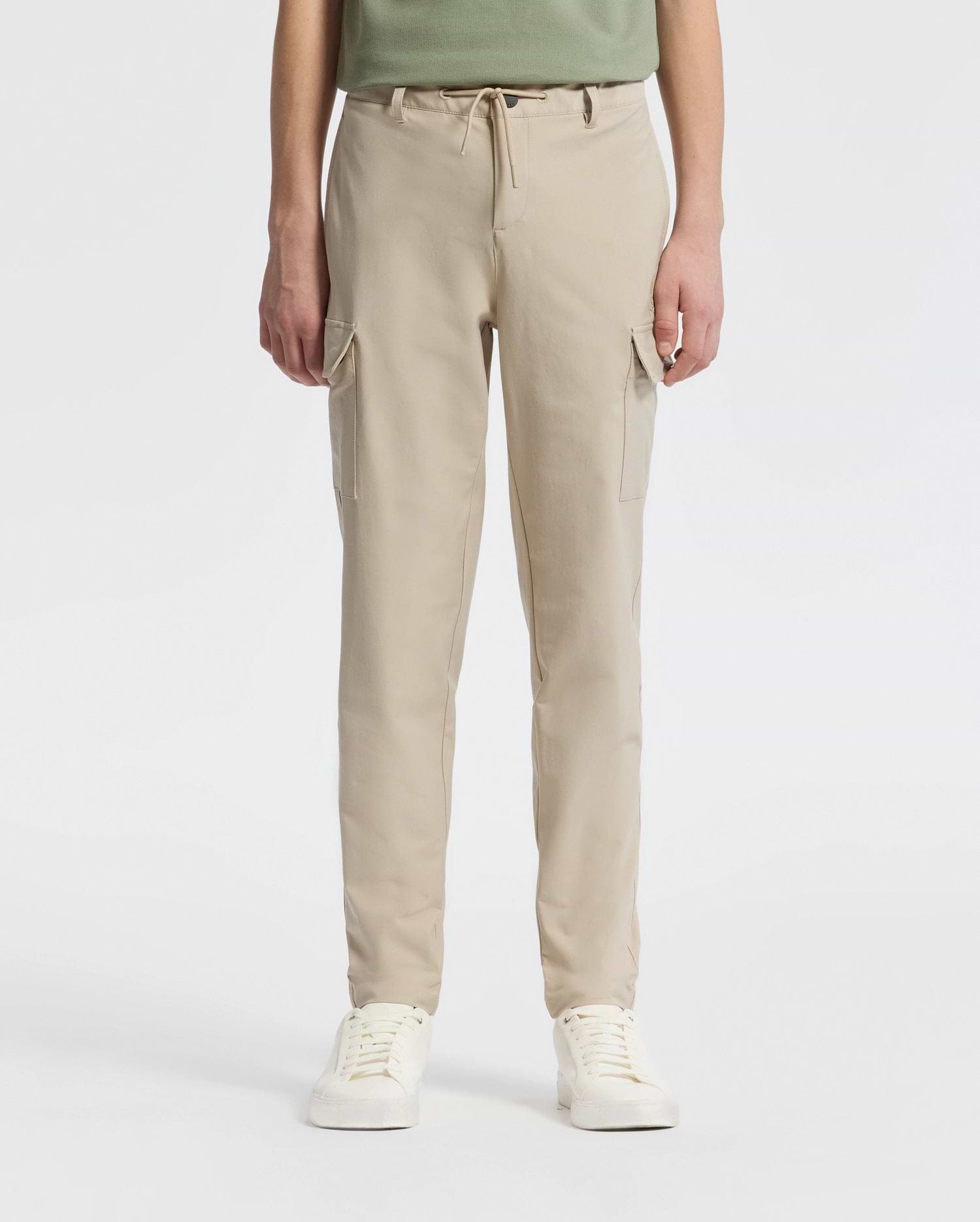 A person stands against a plain backdrop wearing Psycho Bunny’s KIDS GREEN VALLEY CARGO PANT - B0P199E200, featuring beige drawstring waist and multiple pockets, paired with white sneakers. The upper body is partially visible.