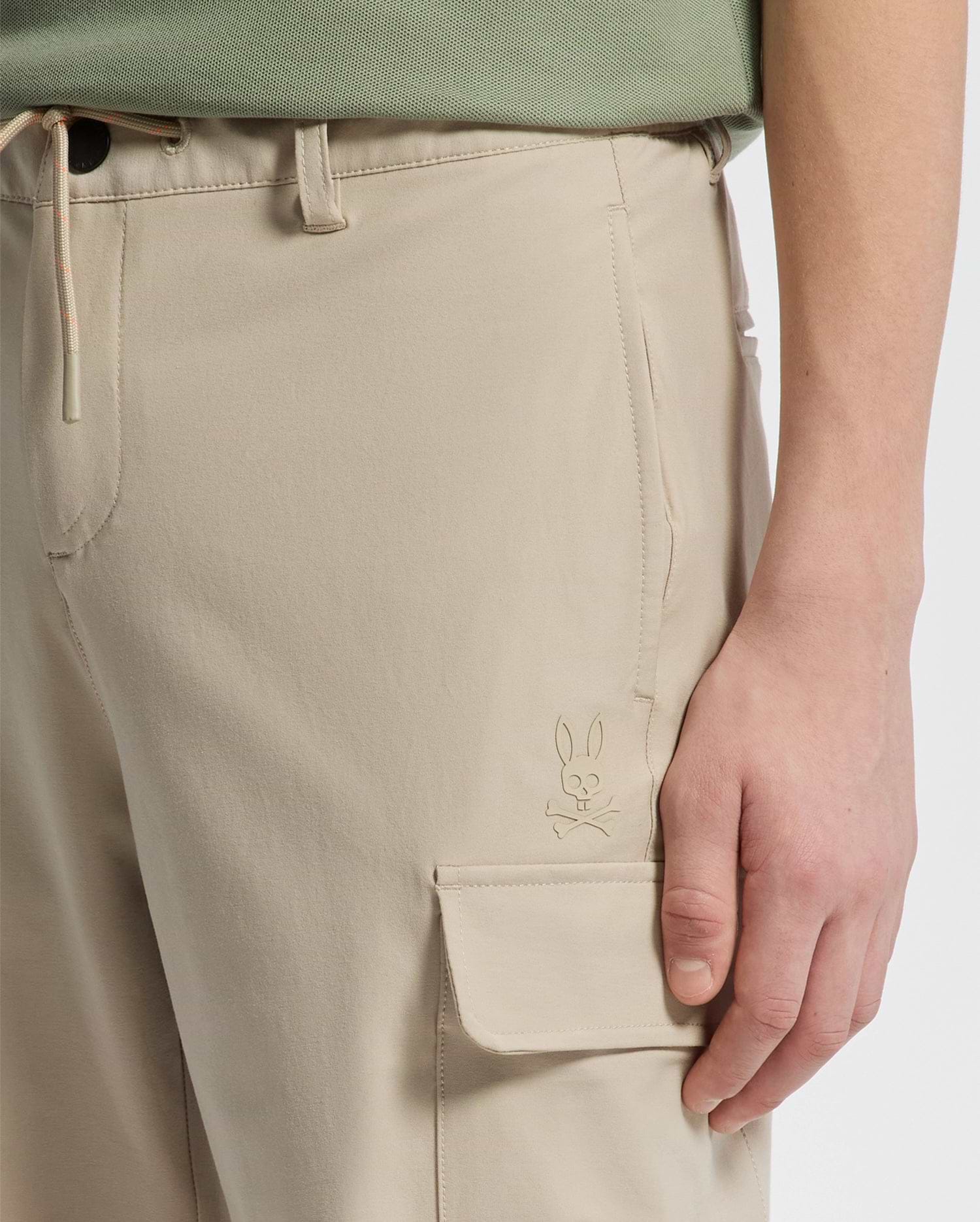 A person wearing Psycho Bunny's KIDS GREEN VALLEY CARGO PANT - B0P199E200, complete with a practical pocket featuring a small rabbit logo. The hand rests nearby, highlighting its design against a plain white background.