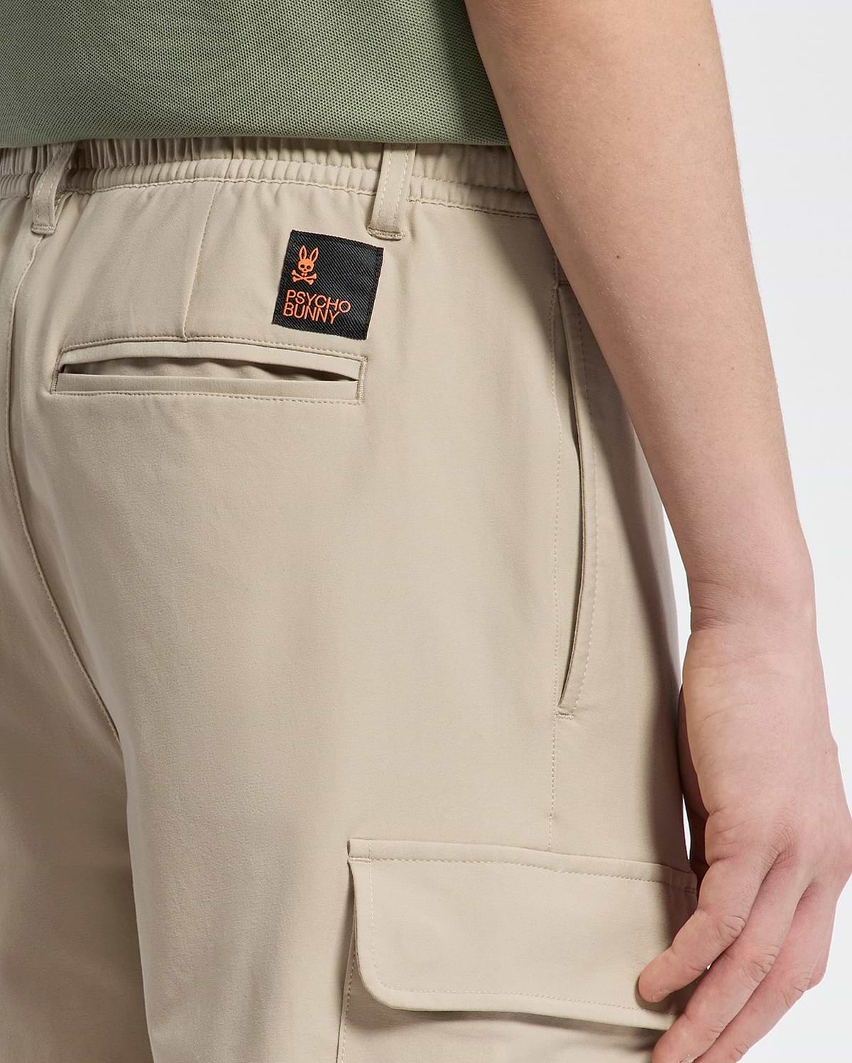 A person is shown from the side in Psycho Bunny's KIDS GREEN VALLEY CARGO PANT (B0P199E200) featuring beige fabric, practical pockets, and an embroidered patch with a small orange design on the back. Paired with a green shirt, this look combines style and comfort seamlessly.