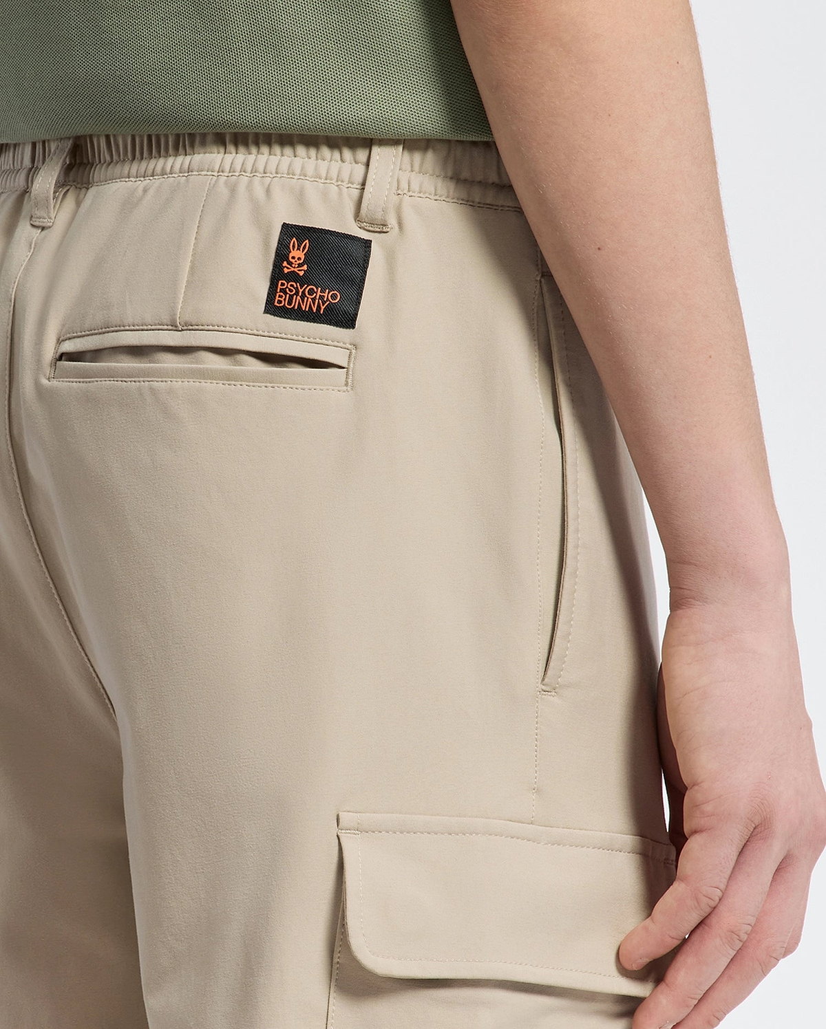 A person is shown from the side in Psycho Bunny's KIDS GREEN VALLEY CARGO PANT (B0P199E200) featuring beige fabric, practical pockets, and an embroidered patch with a small orange design on the back. Paired with a green shirt, this look combines style and comfort seamlessly.