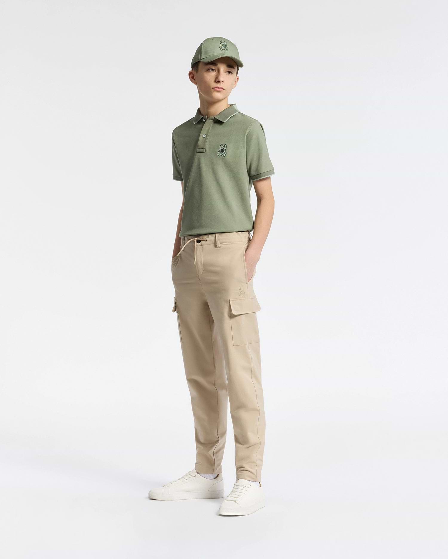 A person wearing a green polo shirt, matching cap, Psycho Bunny KIDS GREEN VALLEY CARGO PANT - B0P199E200 with ample pockets, and white sneakers stands against a plain white background. One hand is in their pocket as they appear comfortably relaxed.