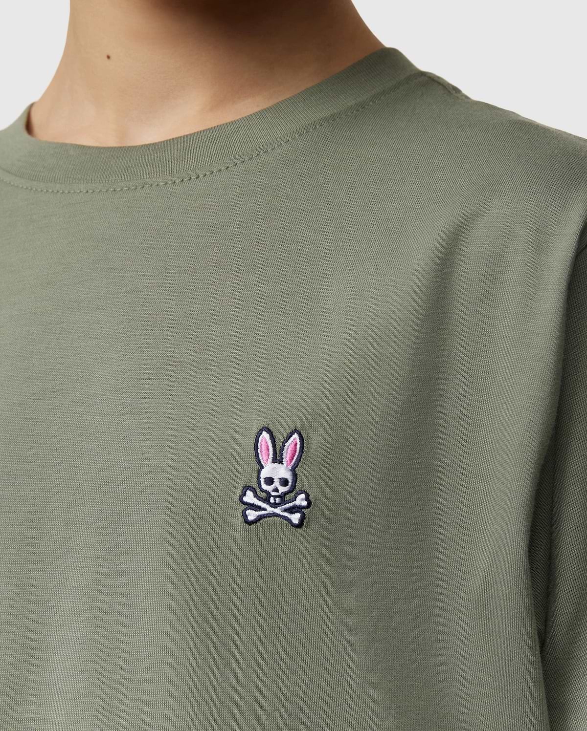 Close-up of a kid wearing the Psycho Bunny Kids Classic Crew Neck Tee in green. Perfect for back-to-school, this 100% Pima cotton tee features an embroidered bunny-headed skull and crossbones logo on the chest.