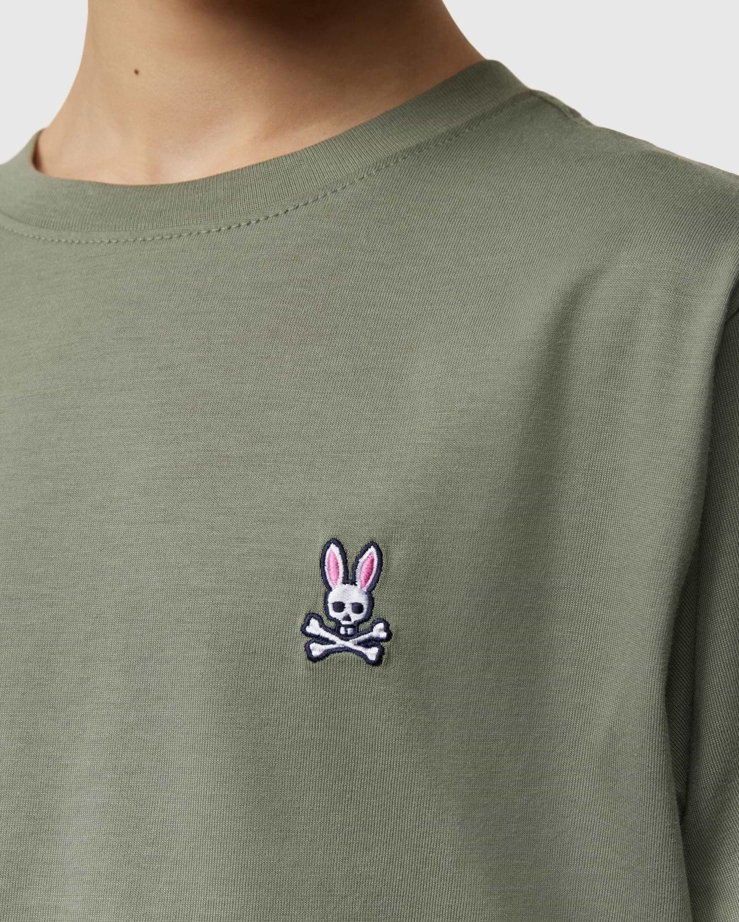 Close-up of a kid wearing the Psycho Bunny Kids Classic Crew Neck Tee in green. Perfect for back-to-school, this 100% Pima cotton tee features an embroidered bunny-headed skull and crossbones logo on the chest.