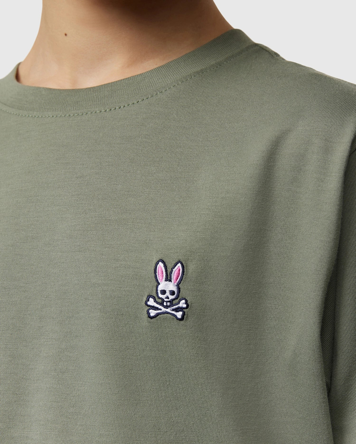Close-up of a kid wearing the Psycho Bunny Kids Classic Crew Neck Tee in green. Perfect for back-to-school, this 100% Pima cotton tee features an embroidered bunny-headed skull and crossbones logo on the chest.