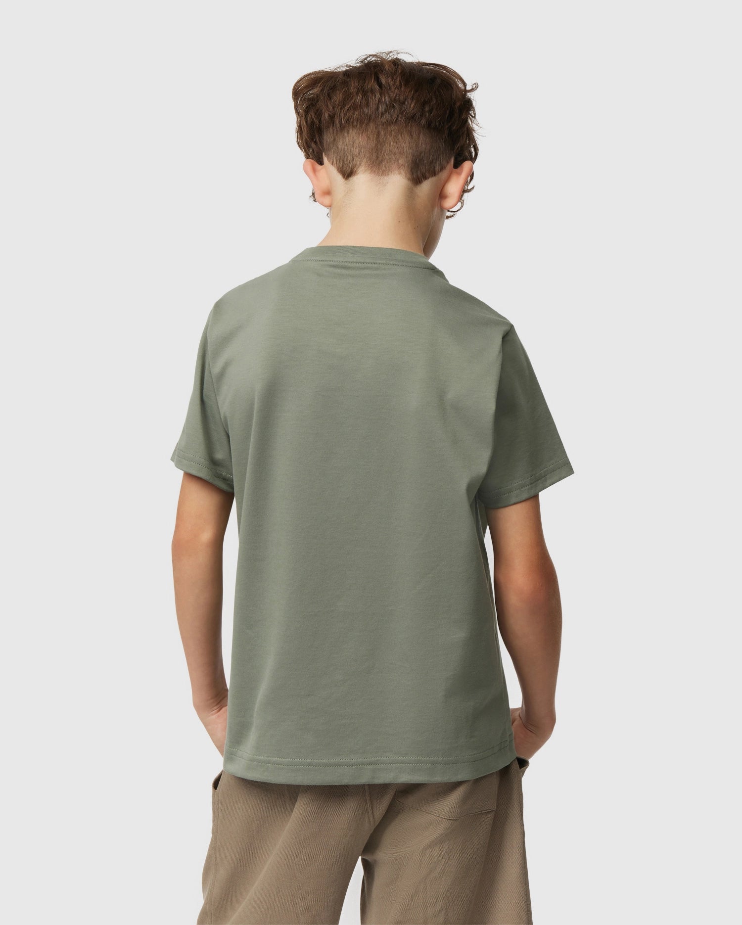 A young boy with short brown hair faces away, wearing a Psycho Bunny Kids Classic Crew Neck Tee in plain green and beige shorts. This 100% Pima cotton tee is ideal for back-to-school, set against a light grey background.