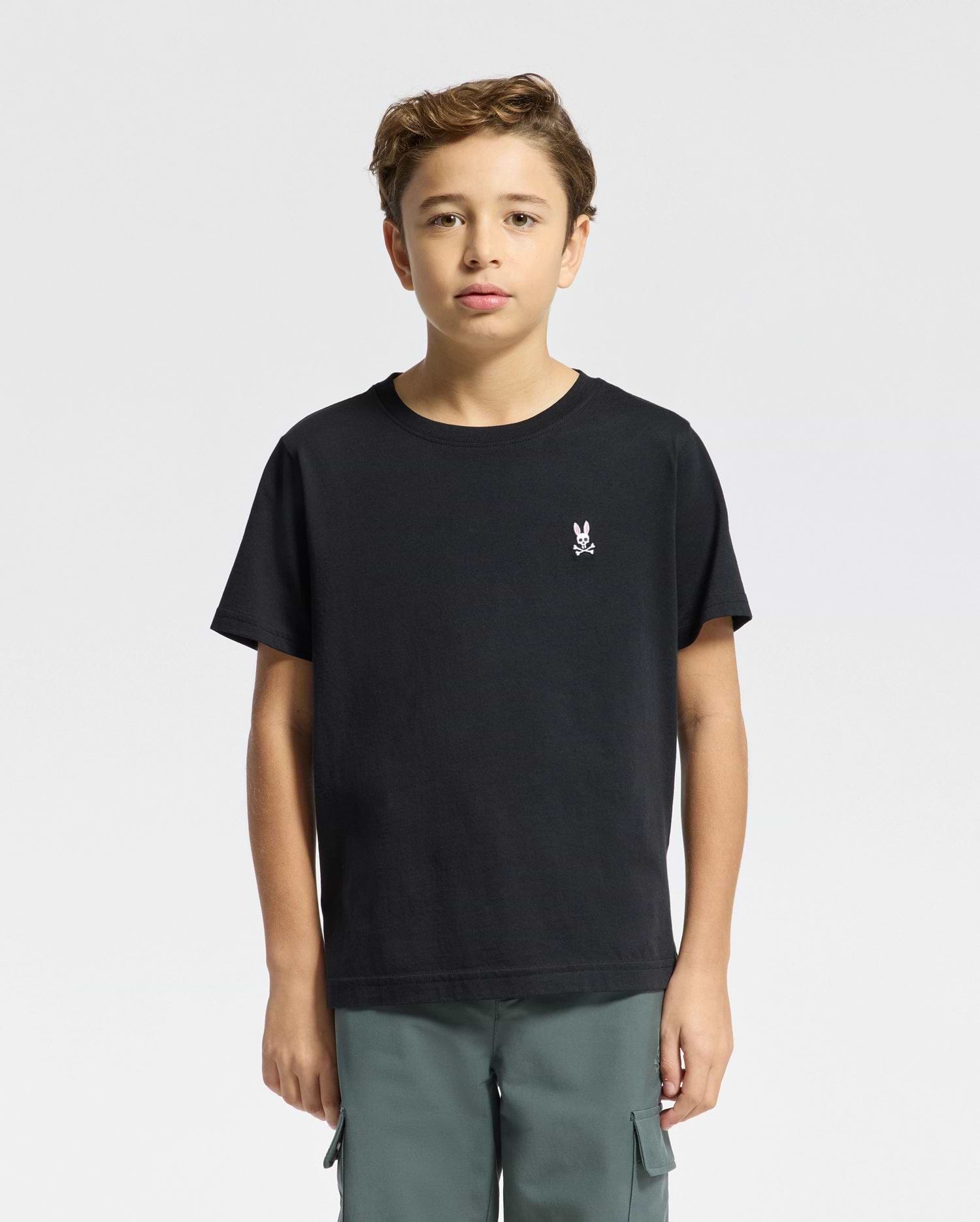 A young person with brown hair is wearing a Psycho Bunny KIDS CLASSIC CREW NECK TEE, made of 100% Pima cotton, showing a small white skull and bunny logo on the left chest. They pair it with green cargo pants against a plain white background.