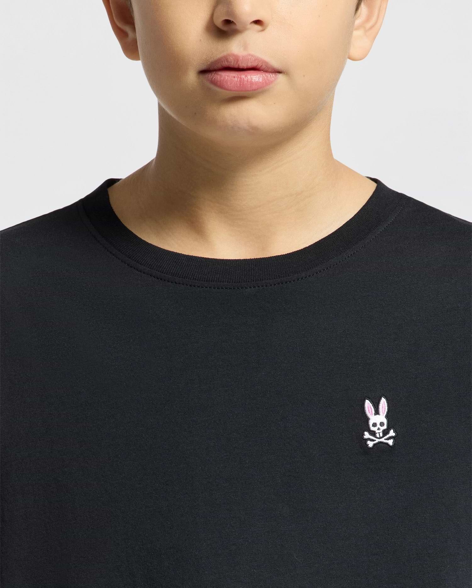 A person wearing the Psycho Bunny KIDS CLASSIC CREW NECK TEE - B0U014CRPC in classic black, made from 100% Pima cotton, features a chest logo with a pink-eared rabbit and crossbones on a plain light-colored background.