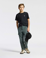 A young boy stands against a plain background, wearing a Psycho Bunny KIDS CLASSIC CREW NECK TEE - B0U014CRPC. His black tee is 100% Pima cotton and pairs with green pants and sneakers. Holding a black cap in one hand, he gazes slightly to the side.