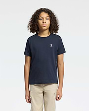 A curly-haired person wears a Psycho Bunny Kids Classic Crew Neck Tee in plain navy, made from 100% Pima cotton with a small chest embroidery. Paired with beige pants against a white background, they represent timeless basics.